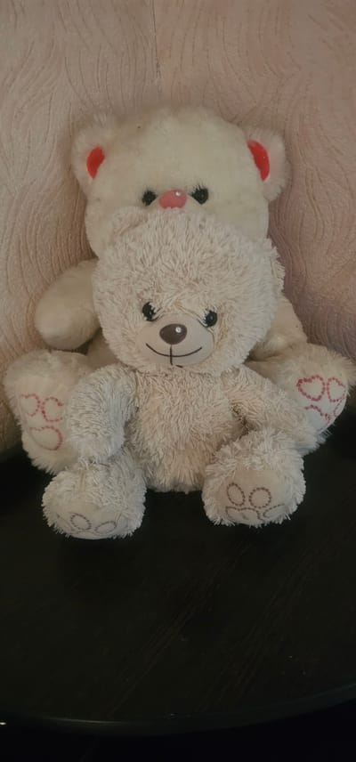 Two white teddy bears are positioned in the foreground and background, seated on a dark, textured surface. The front teddy bear is closer and larger, with a fluffy, off-white fur coat and a brown nose with a light brown embroidered smile and a small, circular, gem-like decoration in the center of its snout. Its eyes are black, round, and shiny. It has red heart shapes embroidered on its paws. The second teddy bear, behind the first, is larger and less detailed, with a more uniform white appearance and pink inner ears. Its nose is pink and its eyes are black. The background is a light-colored, textured wall, possibly wallpaper. The image is taken from a medium-close shot, focusing on the two stuffed toys. The location in Oktiabrskaya, Russia is not discernible from the image.