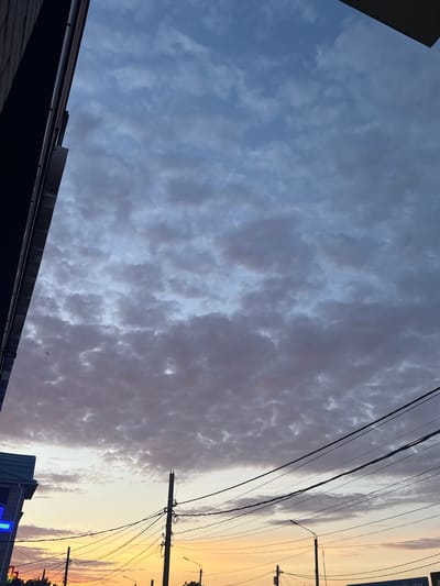 This is a photograph of a sky with dramatic clouds, likely taken at sunset or sunrise. The sky transitions from a pale blue with scattered, textured clouds on the left to a warm, golden hue with wispy clouds on the right. A few dark, silhouetted buildings and rooftops are visible at the bottom of the frame, suggesting an urban or semi-urban setting. Power lines and utility poles stretch across the upper portion of the image, indicating the presence of infrastructure. The overall mood is serene and atmospheric, with the warm tones of the sky creating a sense of peace. The image appears to be taken from a low angle, looking upwards towards the sky. There is no visible text in the image.