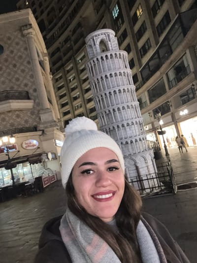 The image displays a nighttime urban scene in Esenler, Turkey. In the immediate foreground, a female individual with dark hair, wearing a white knit beanie with a pom-pom and a grey patterned scarf, smiles directly towards the camera. Behind her, a prominent, large-scale white replica of the Leaning Tower of Pisa stands slightly right of center, its base partially obscured by a dark metal barrier. To the left, a multi-story building features a decorative column and a patterned facade. Below this building, a commercial establishment identified by the illuminated sign "Faruk Güllüoğlu" is visible, featuring glass display cases and outdoor seating arrangements. To the far right, a tall building with multiple illuminated windows indicates residential or commercial activity. On a paved street or pedestrian area extending into the background, two indistinct human figures are visible. The scene is illuminated by artificial light sources from streetlights, building interiors, and commercial signage.