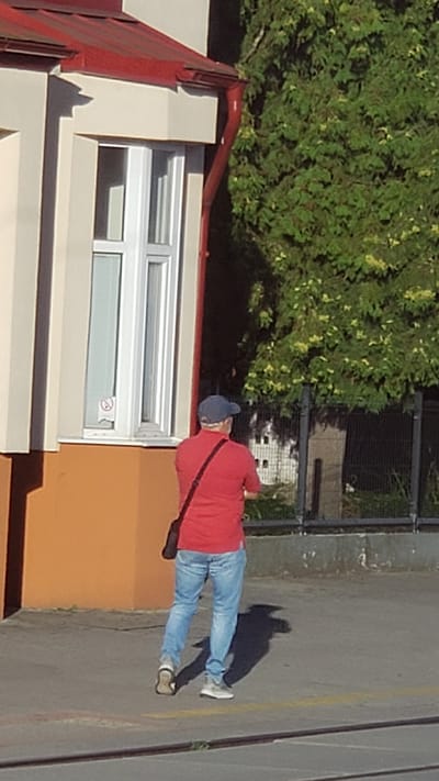 A man wearing a red polo shirt, blue jeans, and a blue baseball cap walks away from the camera. He is carrying a dark bag slung across his chest. The man is in front of a light-colored building with a red roof and gutters. A window on the building has a "no smoking" sticker. The man is walking on a paved surface with a yellow line painted on it, and a fence with foliage behind it. The scene is outdoors in what appears to be an urban or suburban environment. This is the city of Spišská Nová Ves, Slovakia.