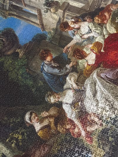 This is a photograph of a jigsaw puzzle depicting a pastoral scene with several figures. The puzzle is laid out on a flat surface, and the image shows a group of people, possibly engaged in leisure activities, in an outdoor setting. There are trees and foliage in the background, and the sky is visible. The figures appear to be dressed in historical or theatrical costumes, with one person playing a stringed instrument and others gathered around. The puzzle itself is composed of numerous interlocking pieces, with the cut lines clearly visible. The overall impression is one of a classical or Rococo-style painting. The photograph is taken from a slightly overhead angle. The image does not contain any elements that would indicate a specific location in Şişli, Turkey.