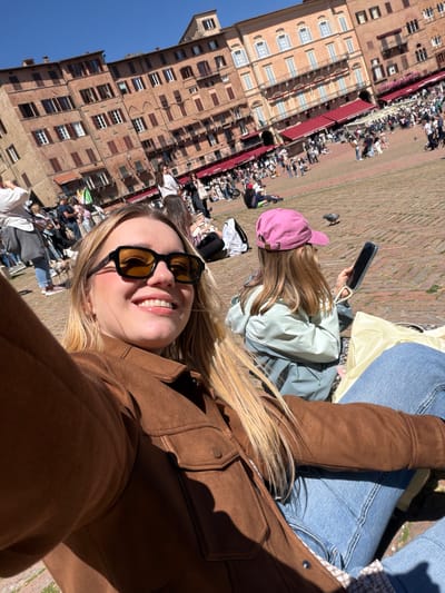 A selfie of a blonde-haired woman wearing a brown jacket and sunglasses, seated in Piazza del Campo in Siena, Italy. In the background is a large crowd of people occupying the piazza, with the Palazzo Pubblico visible. Another woman wearing a pink hat sits nearby, using a smartphone. The piazza is paved with red bricks, and the sky is clear blue.