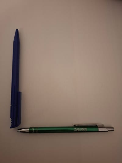 Two pens are arranged in an 'L' shape on a light-colored, textured surface. The vertical pen is blue and appears to be a marker or a large ballpoint pen with its cap on. The cap has a serrated edge. The horizontal pen is green with silver accents and writing that reads "ŽALGIRIS". The pens are positioned in the lower-left quadrant of the image. There is no indication of the location being in the city of Huntingdonshire, United Kingdom.
