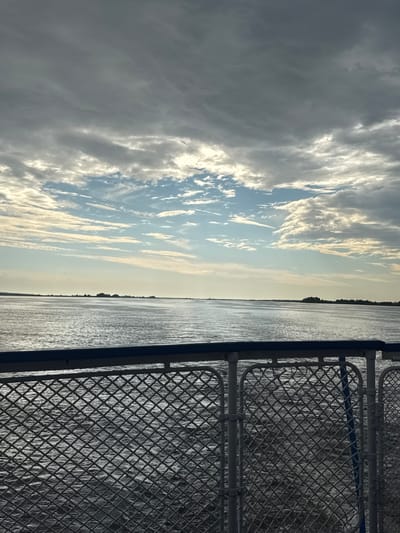 The image shows a view from a vessel, looking out over a body of water towards a distant shoreline. The sky is filled with dramatic clouds, with patches of blue sky and sunlight breaking through. The water is calm and reflects the sky. In the foreground, there is a metal railing and mesh fence, typical of a ship or ferry. The shoreline in the distance appears to be low-lying with some trees and structures. The overall atmosphere is serene and evokes a sense of travel and open space. The image does not contain any discernible elements that would specifically place it in Zuevo, Russia.
