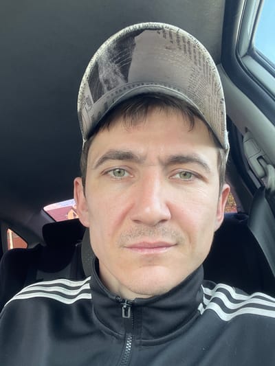 A person is taking a selfie inside a car. The person is wearing a black Adidas jacket with white stripes and a hat with newspaper print. The person has green eyes and stubble. The scene is lit by natural light, suggesting it is daytime. The background shows a car's interior and a glimpse of trees and buildings outside, indicating a suburban or rural setting. The person's expression is neutral. The image contains visible text on the hat, which appears to be in English. The location is identified as Сокуры, Russia.
