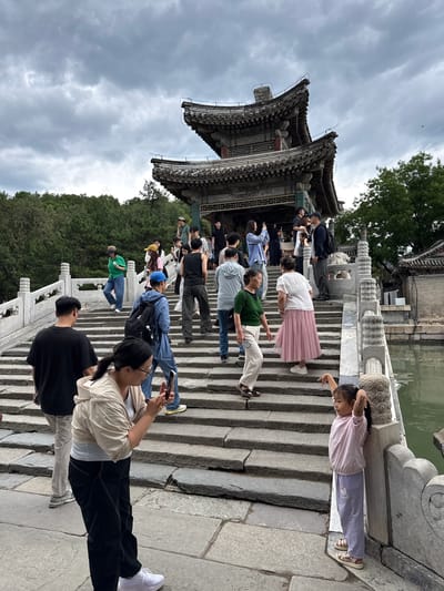 The image depicts a bustling outdoor scene at a multi-tiered traditional Chinese pavilion situated atop a wide stone bridge, likely within the Summer Palace (Yiheyuan) in Haidian District, China. A crowd of approximately 25-30 individuals, predominantly East Asian adults and several children, are ascending and descending the grey stone staircase that forms the bridge. The stairs feature ornate white stone balustrades with decorative pillars.
To the right of the bridge, a tranquil body of greenish water is visible, bordered by another stone structure. Dense green foliage and trees line the background on both sides, suggesting a natural park-like environment. The pavilion itself exhibits traditional Chinese architecture with multiple dark grey tiled roofs, upturned eaves, and visible wooden support structures. The sky above is heavily overcast with a uniform grey cloud cover.
In the immediate foreground, a woman with glasses and a ponytail, wearing a beige hoodie, light grey top, and black pants with white stitching, is holding a smartphone horizontally, appearing to capture media. To her right, a young girl in a light pink long-sleeved top and light purple pants leans against a bridge balustrade, striking a pose with both arms raised and bent at the elbows, looking towards the right side of the frame. Other individuals throughout the scene are walking, conversing, or observing their surroundings; several are holding smartphones, suggesting photography or communication. Clothing is casual, including jeans, t-shirts, hoodies, and comfortable footwear.
