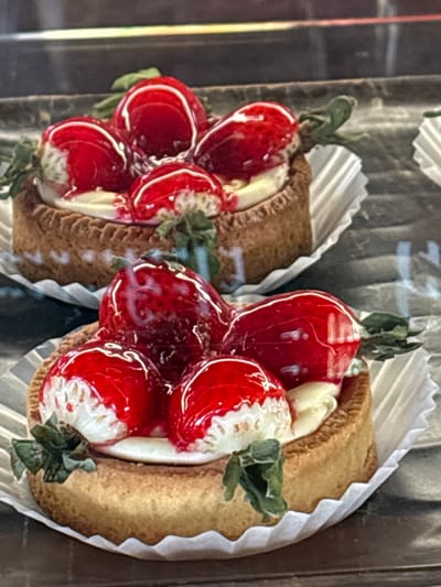 The image displays two strawberry tarts in a retail display setting within Tsovagyugh, Armenia. The foreground tart is in sharp focus, while a second tart is partially visible and slightly blurred in the background. Each tart features a golden-brown, fluted pastry shell filled with a light, creamy substance. Multiple whole and halved red strawberries, coated in a glossy, translucent red glaze, are arranged atop the cream. Small green leaf garnishes are positioned around the strawberries. Both tarts are presented in white, corrugated paper liners and rest on a dark, reflective surface, likely the shelf of a display case.