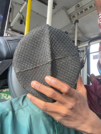 The scene depicts the interior of a public transport vehicle in Ebute-Metta, Nigeria. In the foreground, a person's right hand, with a brown skin tone, holds a dark grey, circular cap directly in front of their face. The cap displays a textured, woven pattern and is stitched into four distinct quadrants. The individual wears a teal-colored garment.
To the subject's left, a black duffel-style bag with a visible zipper and strap rests on a dark grey seatback. Below the bag, a green and white patterned fabric is visible. The vehicle's interior background features white ceiling panels containing horizontal vents, white and yellow grab poles, and a small, dark dome-shaped security camera mounted overhead. A red "STOP" button is affixed to an overhead panel. To the right, another individual is partially visible, dressed in a dark red garment with patterned fabric on the shoulder. A "No Smoking" pictogram is visible on a panel or window frame to the far right, where bright ambient light enters the vehicle.