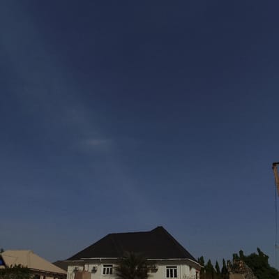 This image depicts a daytime scene in Sámara, Costa Rica, characterized by a predominantly clear, deep blue sky occupying the upper two-thirds of the frame. Minimal, wispy cloud formations are faintly visible in the upper and mid-left areas. In the lower portion of the image, the foreground features architectural structures and vegetation. A two-story building with white facades and a dark, hip-style roof is centrally located. Multiple white-framed windows are visible on its front, along with external air conditioning units. A palm tree partially obscures the lower section of this building. To the far left, a section of another building's light-colored, possibly tan or beige, hip roof is visible. Along the right edge, green foliage and trees are present, partially obscuring a decorative element or statue with a golden hue. A tall, light-brown utility pole or mast is partially visible on the extreme right, with dark cables extending downwards. No individuals are present, and no active actions or interactions are depicted within the frame.