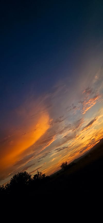 The image depicts a dramatic sunset over the town of Momin Prohod, Bulgaria. The sky is a gradient of deep blue at the top, transitioning to vibrant oranges, pinks, and yellows closer to the horizon. Wispy clouds are illuminated by the setting sun, creating streaks of color across the sky. Silhouetted against this colorful backdrop are the dark shapes of trees and rolling hills, indicating a rural or semi-rural landscape. The overall impression is one of natural beauty and tranquility at the end of the day.