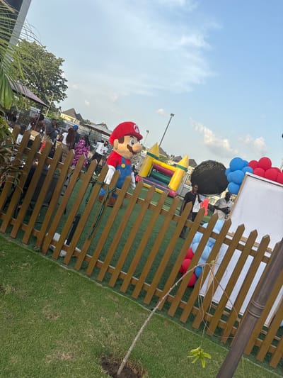 The image depicts an outdoor event in Rubuchi, Nigeria, evidenced by the presence of local residents and architecture. A character resembling Mario from the Super Mario franchise is seen standing next to a wooden fence. Behind the fence, there is a crowd of people, a colorful inflatable bouncy castle, and blue and red balloons. A white sign is also visible in the background. The environment consists of a grassy lawn, a young tree sapling, and some foliage in the foreground. The sky is partly cloudy, suggesting a daytime event.