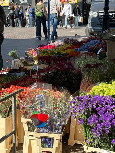 The image depicts a flower stall on a street, likely in Utrecht, Netherlands, given the presence of a McDonald's restaurant. The stall displays a vibrant array of flowers of various colors including purple, yellow, red, and blue, arranged in buckets or containers, some wrapped in clear plastic. People are walking along the sidewalk beside the stall, suggesting it is located in a pedestrian area. A parked car is visible along with a street lamp or sign post.