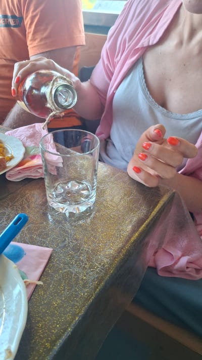 An overhead shot shows a person pouring amber liquid from a glass bottle into a clear, empty glass on a table. The bottle is tied with twine around its neck. The person's hands, with bright orange nail polish, are visible as they hold the bottle and glass. A pink cardigan is draped over the person's shoulder, and they are wearing a light gray tank top. The table surface is covered with a gold glittery tablecloth. To the left, a plate with food and a pink napkin are partially visible. In the background, another person in an orange shirt is seen. The image appears to be taken indoors, possibly in a restaurant or dining area. The location is identified as Дзямідавіцкі сельскі Савет, Belarus.