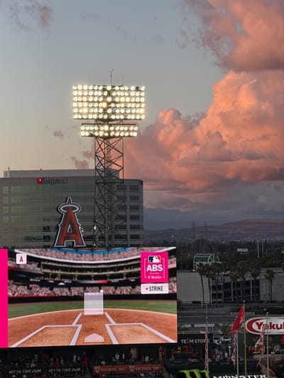 This media file captures an atmospheric evening view of a baseball stadium and its surroundings in Anaheim, United States, likely during an Angels game. The scene is dominated by a dramatic sky at dusk, featuring soft blue and pink hues blending into a vibrant orange as large, fluffy clouds catch the light of the setting sun.

In the foreground, a prominent stadium light tower stands tall, its numerous bright lamps illuminated, casting a warm glow. Behind it, a modern building with the "impact" logo is visible. A large, iconic red "A" logo, representing the Los Angeles Angels baseball team, is prominently displayed on another building, integrated into the stadium complex.

The lower half of the image features a large stadium scoreboard or jumbotron. It displays a graphic of a baseball field from the catcher's perspective, showing a pitch in motion. Text on the scoreboard reads "ABS powered by T-Mobile" and "• STRIKE", indicating an automated ball-strike system or a game replay feature. Below the scoreboard, actual stadium seating areas are visible, with small figures of spectators in the stands. Various advertisements are also visible on the stadium structures, including "NITTO TIRE.COM" and "SALINAS, INC.".

In the mid-ground and background, the landscape includes other buildings, clusters of palm trees, and distant hills or mountains under the colorful sky. Towards the bottom right, flags or banners with the "Yakult" logo and a partial "Monster" (Energy Drink) logo are seen, along with a sign that appears to say "TEAM STOPE" (likely "TEAM STORE"). The overall impression is one of an active evening at a major league baseball venue, enhanced by the picturesque sunset.