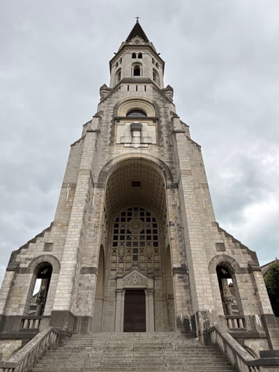 The image shows the facade of a large, stone church, viewed from a low angle looking upwards. The church features a prominent tower topped with a pointed spire and a cross. The facade is ornate, with arched entrances and windows. A wide set of stone stairs leads up to the main entrance. The sky is overcast with clouds. The church is likely the Basilique de la Visitation in Annecy, France, given its architectural style.