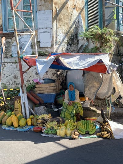 The image displays a street vendor's fruit stall in Lawang, Indonesia, positioned on a paved road against a weathered building facade. An elderly woman is seated centrally beneath an improvised canopy constructed from rusted metal poles and sections of red, white, and blue tarpaulins. She is dressed in a light-colored long-sleeved top, a green and yellow patterned sarong, and a light blue headscarf, with her hands clasped in front of her. On the ground directly before her, a variety of fruits are arranged, including several large bunches of green, yellow, and brown bananas, numerous papayas, green spherical fruits, a small bowl containing red fruit, and assorted root vegetables. Behind the woman, against the stone base and discolored upper wall of the building, are stacked wooden crates, a blue basin, and a metal cabinet. The building's upper facade features a window with peeling white paint on the left and a window with a blue slatted shutter on the right, with multiple weathered papers affixed to the wall. Sunlight illuminates the scene, casting shadows across the foreground pavement.