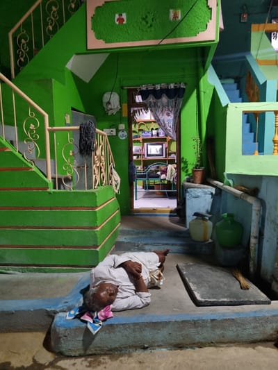 The scene shows an elderly man lying on a concrete step in what appears to be a courtyard or porch adjacent to a building. The man is wearing a white striped shirt and appears to be resting, with a colorful patterned cloth under his head. He is fair-skinned with grey hair. The step he's lying on is painted in blue hues.
The building is vibrantly painted in shades of green and blue, with elements of yellow and peach. The architecture is multi-level, featuring staircases and intricate railings. An open doorway provides a glimpse into the interior of the building, revealing a television, shelves, and furniture, suggesting a living space. The interior is illuminated with what seems like artificial lighting.
Outside the door, various items are visible, including plant pots, brooms, and pipes, adding to the cluttered and domestic atmosphere. The overall impression is of a lived-in space, with a focus on the man taking a rest in the immediate foreground.
The location is Pedda Kammavaripalli, India, based on the geo-tag.