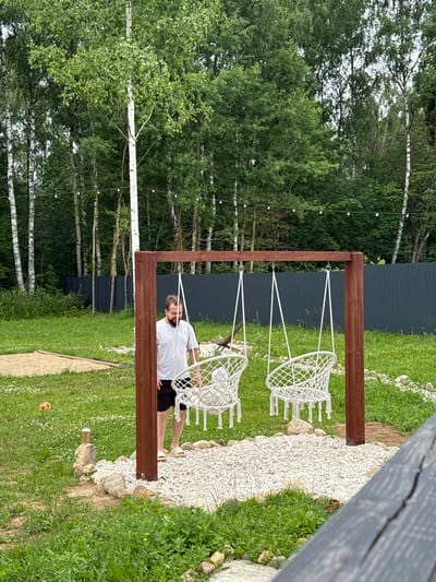 In Ryzhkovo, Russia, a man stands near a wooden swing set in a backyard. The swing set has two white, macrame-style seats hanging from white ropes. The ground beneath the swings is covered with white gravel, surrounded by rocks and green grass. The man, wearing a white t-shirt and black shorts, is looking towards the swings. Behind the swing set, a dark grey corrugated metal fence runs along the property line, backed by a dense line of trees, including birch trees. String lights are visible in the background, stretched between trees. To the left of the swing set, a small orange ball rests on the grass. The overall environment appears to be a residential outdoor space on a cloudy day.
