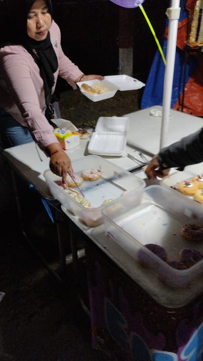 At an outdoor food stall in Sukodono, Indonesia, a woman wearing a pink striped shirt and a black hijab is serving food. She is holding a white styrofoam container filled with rice and a piece of fried food. Next to her, a person's arm is reaching into a clear plastic container filled with donuts. Another clear plastic container to the right also contains donuts. The stall appears to be set up on a table, with various food items and containers visible. The background is dark, suggesting it is nighttime, with a blue tarp and a white pole also present.