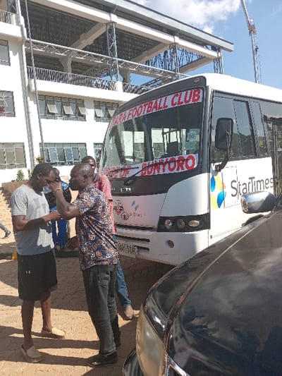 This outdoor image, likely taken in Siaya, Kenya, during a bright, sunny day with a blue sky and scattered white clouds, features a large white passenger bus, several men, and part of a stadium-like structure with a construction crane.
The focal point is a white Tata bus, branded with "StarTimes" on its side. Across the front windshield, two red and white banners are displayed, clearly reading "BUNYORO FOOTBALL CLUB" on the top one and "PRIDE OF BUNYORO" on the bottom. The bus's license plate, "KDE 575F," is also visible. It is parked on a dirt or gravel surface, with a dark-colored car partially visible to its right.
Three men are in the foreground. Two are closer to the camera, standing and appearing to interact casually. One man wears a patterned short-sleeved shirt and dark trousers, while the other wears a grey t-shirt and black shorts. A third man is partially visible further back, near the bus.
In the background, a large, multi-story building with a robust structural framework, resembling a stadium or a large public facility, stands prominently. A tall construction crane is also visible against the sky, suggesting ongoing development or a significant infrastructure project at the location. The scene appears to be a parking area or entrance to this facility.