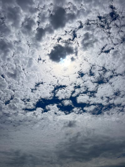 The image is a photograph of a sky filled with a mixture of white and gray clouds. The sun is partially obscured by a darker cloud in the center, creating a luminous halo effect. The clouds are textured and appear to be a type of altocumulus cloud formation, with many small, rounded masses. The overall impression is of a dynamic and somewhat overcast sky, with patches of blue visible between the clouds. The photograph is taken from a low angle, looking upwards. The location is identified as Diepenbeek, Belgium. There are no people, subjects, objects, or actions depicted in the image, as it is solely focused on the sky and cloud formations.