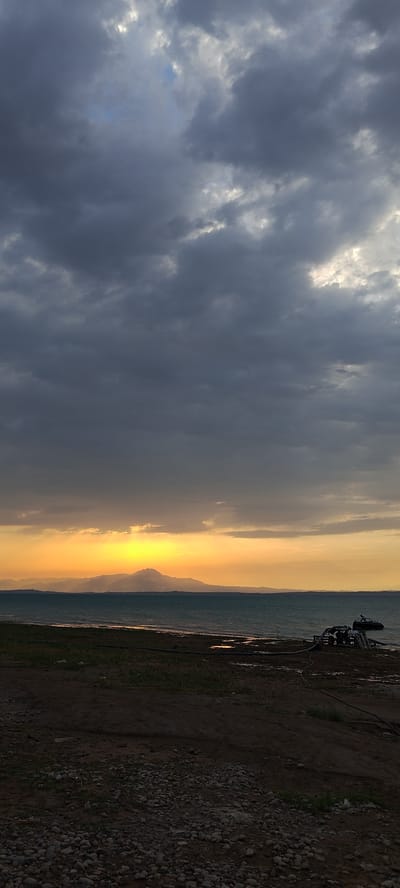 The image depicts a sunset over a body of water, with a mountain range in the distance. The sky is filled with dramatic, dark clouds, with a bright patch of sunlight breaking through on the horizon. The water is choppy, and there is a small, dark object that appears to be a boat or raft on the right side of the frame. The foreground consists of a rough, pebbly ground with some sparse vegetation. The scene is likely set in Chigdalik, Tajikistan, given the mention of the location. There are no people visible in the image.