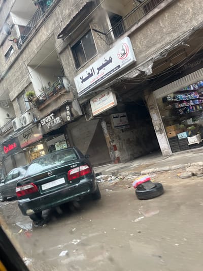 The image depicts a street scene in Jaramana, Syria, during daytime under overcast conditions. A dark green Mazda 626 sedan, bearing Syrian license plate number 844370 Damascus, is parked on the wet pavement in the foreground, with its rear facing the viewer. Another silver sedan is partially visible behind the Mazda to the left. The street surface is uneven, wet with puddles, and littered with various debris and trash. To the right of the Mazda, a discarded tire is positioned on the pavement, topped with a red and white striped object, possibly a cushion or packaging.

The background consists of a weathered multi-story building, combining residential and commercial functions. The ground floor features several storefronts. From left to right, visible signs include "Cana 2", "Stylish", "SPORT WEAR", and multiple signs in Arabic for "مختبر الغلا" (Al-Gala Laboratory), one prominently displayed on an illuminated sign with a medical logo and contact information, and smaller ones below it. One storefront on the far right is open, revealing shelves stocked with various goods and boxes. Above the storefronts, multiple air conditioning units are affixed to the building's facade. Exposed electrical wiring is visible along the building's exterior. Upper floors feature balconies, some adorned with plants. No individuals are present in the scene.
