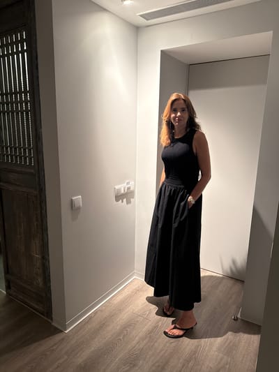 In Grândola, Portugal, a woman stands in a well-lit hallway. She is wearing a black sleeveless dress with a ribbed bodice and a full skirt, complemented by black thong sandals. Her left hand rests in the pocket of her dress, and she wears a watch on her left wrist. The hallway features light gray walls, a light gray baseboard, and a light wood-grained floor. To the left, a dark, intricately carved wooden door is visible. Light fixtures are recessed into the ceiling, and a horizontal vent is present above a doorway. Light switches are mounted on the wall to the woman's left. The overall impression is one of modern architecture with a touch of traditional design.