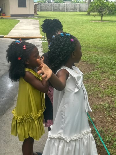 The image shows three young Black girls standing outside, with two of them in the foreground and one partially visible behind them. The girl on the left is wearing a bright yellow dress with ruffled trim at the hem and has her hair styled in braids adorned with colorful hair clips. She is looking to her left and her left hand is raised, appearing to interact with something just out of frame or to be posing. The girl in the center, wearing a white dress with a ruffled hem and a necklace, is looking to her right, with her arm resting on the shoulder of the girl in yellow. The third girl, visible behind the other two, has her hair in braids as well and is facing away from the camera. They are standing on a paved surface next to a grassy area. In the background, there is a light-colored building with a dark window, a concrete wall, lush green grass, and a small tree. The overall setting suggests an outdoor environment, possibly a school or residential compound in Itoga, Nigeria.