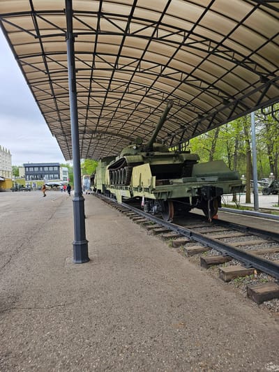 The image shows an army tank on a railroad track under a metal awning, adjacent to a paved walkway. The tank is green and appears to be a static display. The railroad track runs parallel to the walkway. In the background, there are trees, buildings, and some people walking. The photo was taken in Arkhangel'skoye, Russia.