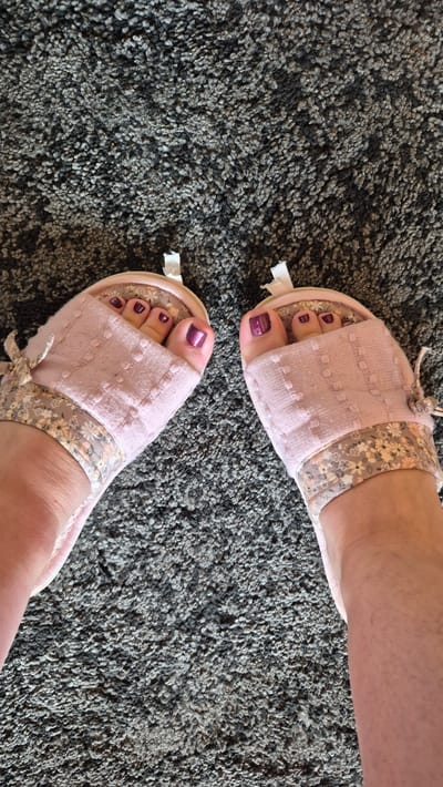 A pair of feet wearing open-toed pink slippers with floral accents are visible against a grey carpeted floor. The toenails are painted a metallic purple. The scene is an indoor environment, possibly a home, and the slippers are positioned centrally on the carpet. The image captures a close-up view of the feet and slippers, with the carpet filling the background. There is no indication of the location being in Krauja, Latvia.