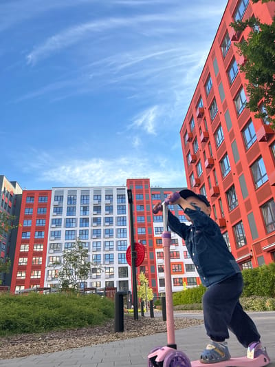A young child rides a pink scooter with purple accents on a paved path. The child is wearing a denim jacket, dark pants, and blue sandals. In the background, there are two modern apartment buildings. One building is primarily white with red accents, and the other is predominantly red with dark gray accents. Both buildings have numerous windows. The sky is bright blue with wispy white clouds. The scene suggests a sunny day, likely late afternoon or early evening given the angle of the light. The setting appears to be a residential area or courtyard in Kaluga, Russia, with greenery visible between the buildings. The child's posture and the overall composition imply an active and playful moment. The word "GRIT" is visible on the scooter's handlebar.