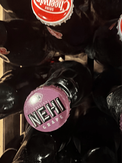 The image shows a close-up view of several glass bottles, presumably soda, packed together. The bottle caps are visible, with one prominently displaying a pink cap labeled "NEHI GRAPE" in white lettering. Another cap is partially visible, with a red background and white "Cheerwine" lettering. The bottles appear to be dark in color. The environment suggests a storage or display setting, possibly a convenience store, in Rochester, United States.