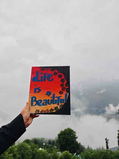 A person's hand holds up a painting with the words "Life is beautiful" written on it, in the city of Prini, India. The painting is predominantly orange and red, with blue lettering and black floral designs. The background is a cloudy sky and a misty mountain landscape with green trees.