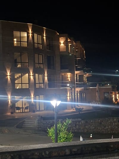 The image shows a modern, multi-story building at night in Tyulenovo, Bulgaria. The building has a facade of textured stone or brick and is illuminated by strategically placed uplights that highlight architectural features and windows. Several windows on the upper floors are lit from within, suggesting occupancy. On the ground floor, large glass doors lead into what appears to be an interior space with furniture visible, including chairs and tables. Outside, there are steps leading up to the building entrance, a stone wall, and landscaping with small trees. A modern lamp post with a bright light is positioned in the foreground, casting a significant glare across the scene. In the background, other lights are visible, and the dark sky suggests the late hour. The overall impression is one of a well-lit, contemporary establishment, possibly a hotel or restaurant, in a coastal or rural setting.