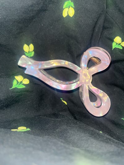 The image displays a pink, iridescent hair claw clip positioned on a black fabric background adorned with a pattern of small yellow flowers with green leaves. The clip has a bow-like shape with two interlocking loops. The fabric appears to be a garment, possibly a dress or skirt, with visible folds and textures. There are no people or any indication of a specific location within Conuco Viejo, Venezuela. The scene is a close-up, still life composition.