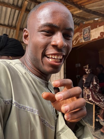 The image depicts a close-up of a young adult male, of apparent African descent, located in Kanti, Nigeria. The subject, positioned in the foreground, has a shaved head, dark skin, and is smiling broadly towards the viewer, revealing both upper and lower teeth. His eyes are slightly narrowed from the smile. He is wearing a light olive-green garment, possibly a tunic or kaftan, with distinctive purple geometric embroidery along the neckline and chest. His right hand is partially visible in the lower right, with fingers slightly curled forward, possibly mid-gesture.
In the background, on the left, a person with dark skin and a black head covering is partially visible. Further in the background, to the right of the main subject, a structure with a corrugated metal roof and wooden supports is discernible. Another individual, also of apparent African descent, is seated or standing in the middle ground on the right, wearing a patterned garment featuring geometric designs in brown, beige, and black. A dark brown wall or surface behind this individual exhibits painted decorations, including circular motifs and abstract patterns. Additional indistinct figures are present in the deeper background. The scene is brightly lit, indicative of daylight conditions.
