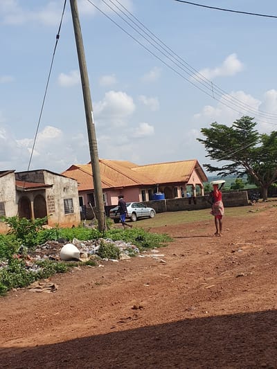 The image captures a vibrant, sunlit street scene in what appears to be a rural or semi-urban area of Idah, Nigeria. The dominant feature is a wide, unpaved dirt road, characterized by its reddish-brown earth, which stretches diagonally across the frame from the foreground towards the background.
In the middle ground, a woman walks barefoot along the dirt path, carrying a large, flat, light-colored object expertly balanced on her head. She is dressed in a red top and a colorful patterned skirt. Behind her, a large tree with lush green foliage provides shade, and a substantial building with a reddish-orange corrugated roof and light-colored walls is visible. A silver car is parked near this building, with a person standing close by.
To the left of the road, a prominent utility pole rises into the bright blue sky, carrying multiple power lines that crisscross the upper left quadrant of the image. Further down, closer to the foreground, is another, older-looking building with a grey, weathered facade. Along the edge of the dirt road, to the left, there's a strip of green vegetation intertwined with a considerable amount of scattered trash and debris, including plastic bottles and other discarded items. An overturned white pot lies amidst this refuse.
The sky is a clear blue with scattered white, cumulus clouds. The strong sunlight indicates it is daytime, likely mid-morning or afternoon, casting visible shadows on the ground. In the distance, beyond the buildings and trees, hints of green hills or a natural landscape can be seen. No clear, readable text is visible on any objects within the image.