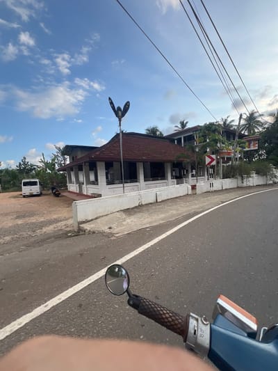 The image captures a roadside scene near Angulugaha Junction, Sri Lanka. The view is taken from the perspective of someone on a moving vehicle, likely a scooter, as part of the handlebar and a side mirror are visible on the right side of the frame.
To the left, a two-story building with a red tile roof stands parallel to the road. A white wall runs along the edge of the building's property, bordered by a narrow dirt area and the road. A white van is parked in the dirt area near the building, slightly further down the road. A three-armed street light fixture is also located by the building.
The road itself is asphalt with a white dividing line. Palm trees and other greenery can be seen in the background, suggesting a tropical environment. Power lines stretch across the top left portion of the image against a partly cloudy sky. The overall scene appears to be a typical roadside setting in a semi-urban or rural area of Sri Lanka.