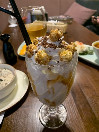A close-up, eye-level, indoor shot captures a dessert in a stemmed glass, topped with whipped cream, chocolate sprinkles, and caramel-coated popcorn. The glass is filled with a creamy, white substance with visible caramel swirls. Two black straws are inserted into the dessert. To the left of the dessert, a white bowl with a creamy soup containing dark specks is visible, placed on a saucer. Further back and to the left, a dark bottle with a spout sits on the table. Behind the dessert, a partially filled glass of amber liquid and a white bowl with food are discernable. The setting appears to be a restaurant in Severodvinsk, Russia, with a wooden table surface. The lighting is dim, suggesting an evening or indoor ambiance. No individuals are clearly visible in the frame.