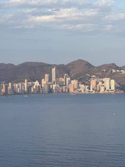 This is a vertically oriented, medium-distance aerial photograph capturing a coastal cityscape bordering a body of water. The scene is dominated by a densely populated urban area nestled against a steep, arid hillside. Numerous multi-story buildings, primarily hotels and apartment complexes, are packed tightly together along the coastline, creating a striking contrast with the natural landscape. Some buildings are white, others are a pale beige, and a few exhibit colorful facades, including a prominent reddish-pink structure.

The buildings are situated on a relatively narrow strip of land between the sea and the mountain. The hillside is sparsely vegetated with scrub and appears rocky. The sea stretches out to the right of the frame, appearing calm with gentle ripples and a subtle texture from the wind. There are a few small white dots visible on the water, likely boats or buoys. In the upper right portion of the water, a small, yellow object, possibly a buoy or a small boat with a canopy, is visible.

Above the cityscape and mountain, a pale blue sky is visible, partially obscured by wispy white clouds that are elongated and streaked, suggesting a light breeze. The lighting indicates it is likely late afternoon or early evening, as the sun appears to be casting a warm, soft light on the buildings and creating subtle shadows. The overall impression is one of a bustling tourist destination situated in a picturesque Mediterranean setting. No text is visible in the image.