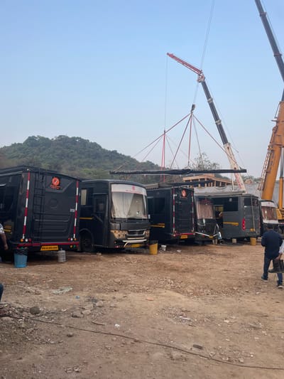 The scene depicts a roadside location in Mira-Bhayander, India, where a construction project appears to be underway. A row of black luxury vans, labeled "SIDOHI VINAYAK VANITY VAN," are parked on a dirt and gravel surface. Overhead, two cranes, one labeled "LAXMI CRANE SERVICE" and "MASTAN CRANE SERVICE", are being used to lift and position wooden beams or structural elements. A tree covered hill can be seen in the background. At least one individual is standing near the vans, while another is possibly directing the crane operation. The vans are parked facing the side of the road with their back bumper lights facing forward. The ground is mostly barren and littered with rocks and debris.