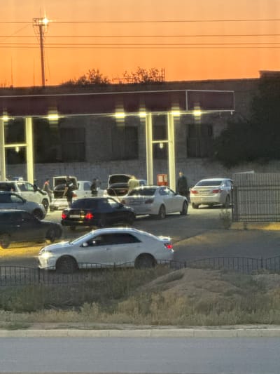 The image depicts a scene with several cars parked, some of which appear to be at a gas station. The time of day is likely dusk or dawn, given the warm orange glow of the sky and the artificial lights illuminating the area. Several people are visible, some standing near the cars, possibly refueling or socializing. One person is leaning into the trunk of a dark-colored car. Another individual, wearing a red hat, is standing near the rear of a white car. The cars are of various models and colors, including white, silver, and dark hues. The setting appears to be an urban or suburban environment, with a building or structure in the background and a grassy area bordering a paved surface. The overall atmosphere seems relaxed, with people engaged in casual activities. There is no visible text in the image.