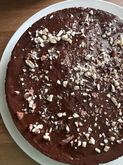 A close-up, overhead shot depicts a chocolate cake on a white plate, presumably indoors. The cake is round and coated in a glossy, dark brown chocolate glaze. It is decorated with chopped nuts, predominantly white, with some darker brown nuts mixed in. The plate sits on a wooden surface. The location is unknown.