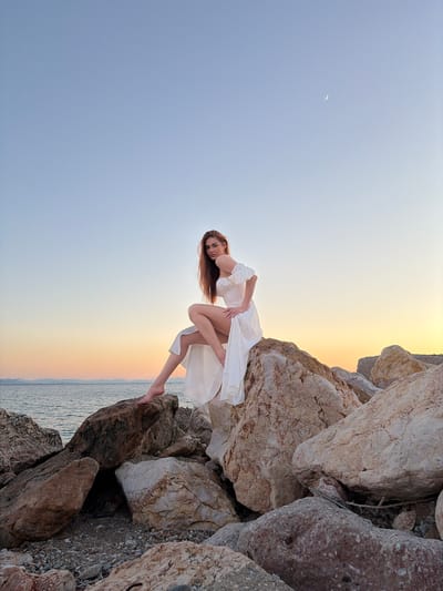 A young woman with long, reddish-brown hair sits on a large rock formation on a rocky shore. She is wearing a white, off-the-shoulder dress with ruffled sleeves and a high slit. Her left leg is bent at the knee, and her bare foot is visible. The ocean is behind her, with a calm body of water and distant hills. The sky is a gradient of pale blue at the top, transitioning to orange and yellow near the horizon, indicating either sunrise or sunset. A faint crescent moon is visible in the upper right portion of the sky. The rock formation she is sitting on is composed of large, irregularly shaped boulders. The foreground is filled with smaller rocks and pebbles. The overall scene is natural and serene, with a sense of soft light and a tranquil atmosphere. The location appears to be a coastal area, possibly a beach or rocky inlet, consistent with the coastline of Paleo Faliro, Greece, known for its seaside promenades and rocky shores.