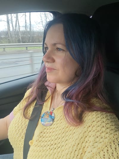 A woman is depicted in profile inside a car, looking contemplatively out the window. She has dark hair with vibrant blue and pink/purple streaks, framing her face. She is wearing a light yellow, open-knit cardigan over a pale pink top, with a seatbelt visible across her chest. A unique, colorful pendant with an abstract design hangs from her neck.

The car's interior is partially visible, including a dark seat and window frame. Outside the window, the scenery appears blurred, indicating motion. Bare deciduous trees, possibly birches, line what looks like a road, separated by a metal guardrail. The sky is overcast, suggesting daytime, perhaps in early spring or late autumn. A faint "Ford" logo can be seen on the car window. The setting is within the context of traveling through Сухарево, Russia.