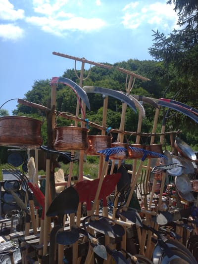 This outdoor shot captures a vibrant display of traditional farming tools and copper cookware, likely at a market or fair, given the abundance and arrangement of items. The scene is set against a backdrop of lush green trees and a bright blue sky with scattered white clouds, suggesting a pleasant, sunny day, possibly late morning or early afternoon.
The foreground is dominated by various tools with wooden handles, including scythes with curved metal blades, shovels with brightly colored red and black heads, and several rakes, some with blue plastic tines. Hanging from wooden racks are several hammered copper pots and kettles, reflecting the sunlight and adding a warm, metallic sheen to the display. In the background, a dense forest covers a hillside, providing a natural and picturesque setting.
The overall impression is one of a bustling marketplace with a focus on handcrafted and traditional items. The variety of tools and cookware suggests a theme related to agriculture, gardening, or possibly historical reenactment. There are no visible people in the image, but the arrangement of goods implies a vendor has set them up for sale. The clear sky and bright sunlight indicate good weather, conducive to an outdoor event. The location context of Ilidža, Bosnia and Herzegovina, hints at a cultural heritage display, possibly showcasing local craftsmanship.