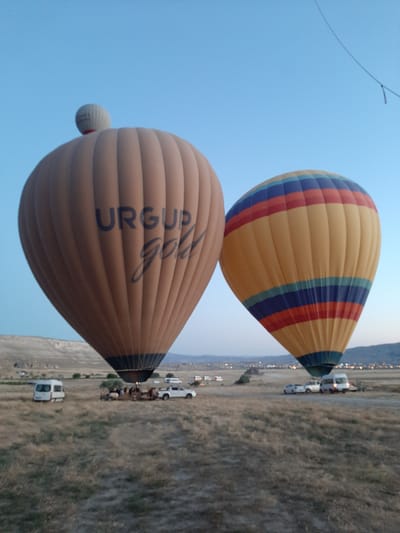 The image depicts a landscape scene with two hot air balloons, one predominantly brown with "URGUP Gold" written on it, and another with yellow, red, and blue stripes. Both balloons appear to be in the process of launching or landing, as evidenced by people gathered near the base of the brown balloon and several vehicles, including a white van and cars, surrounding the landing area. The balloons are situated in a dry, grassy field with rolling hills in the background. The sky is a clear, light blue, indicating it is either early morning or late evening. The overall setting suggests a popular tourist destination known for hot air balloon rides. The landscape and the characteristic geology point to Cappadocia, a region in Turkey, with Avanos being a notable town within this region, known for its pottery and hot air balloon tourism.