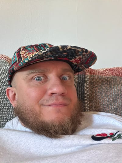A man with light skin, blue eyes, and a beard is looking directly at the camera. He is wearing a colorful patterned hat and a white shirt with an embroidered flower design near the collar. The background includes a textured wall and a patterned cushion. The location is unspecified in the city of Morton Grove, United States of America.
