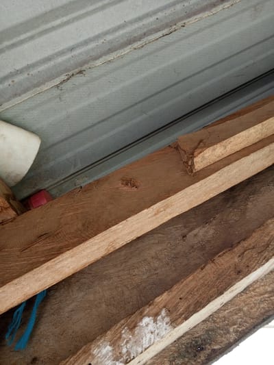 The image shows a close-up view of corrugated metal roofing and wooden planks. The metal roofing is grey and ribbed, with some dirt and small reddish-brown spots on its surface. Below the metal roofing, there are several wooden planks. One plank is lighter in color and appears to be newer, while others are darker and show signs of wear, with visible grain and some white powdery residue on their surface. A piece of bright blue string is hanging from one of the wooden planks. In the upper left portion of the image, a portion of a white cylindrical object is visible, and next to it, a small pink rectangular object is nestled between two wooden planks. The overall scene suggests an unfinished construction or a storage area, likely in an outdoor setting. This image was taken in the city of Gindin Dutse, Nigeria.